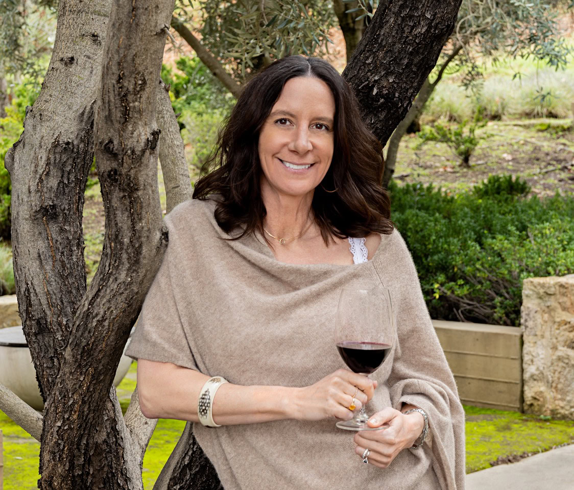 Woman holding a glass of red wine outdoors near trees and greenery.