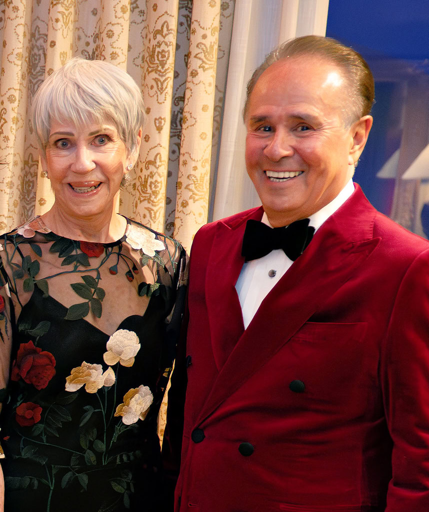 Smiling elderly woman and man in tuxedo at a formal gathering, standing in front of patterned curtains.