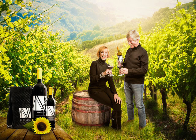 Happy couple enjoying wine in a vineyard with bottles and a barrel in the background.